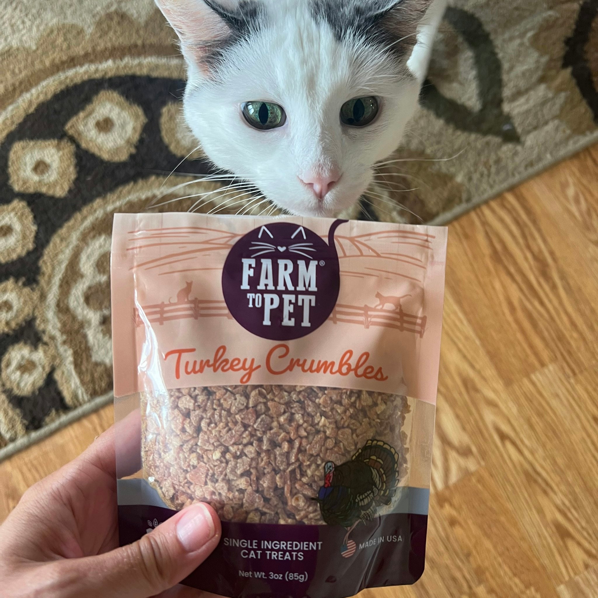 A white and gray cat with a bag of Farmto-pettreats Turkey Crumbles .