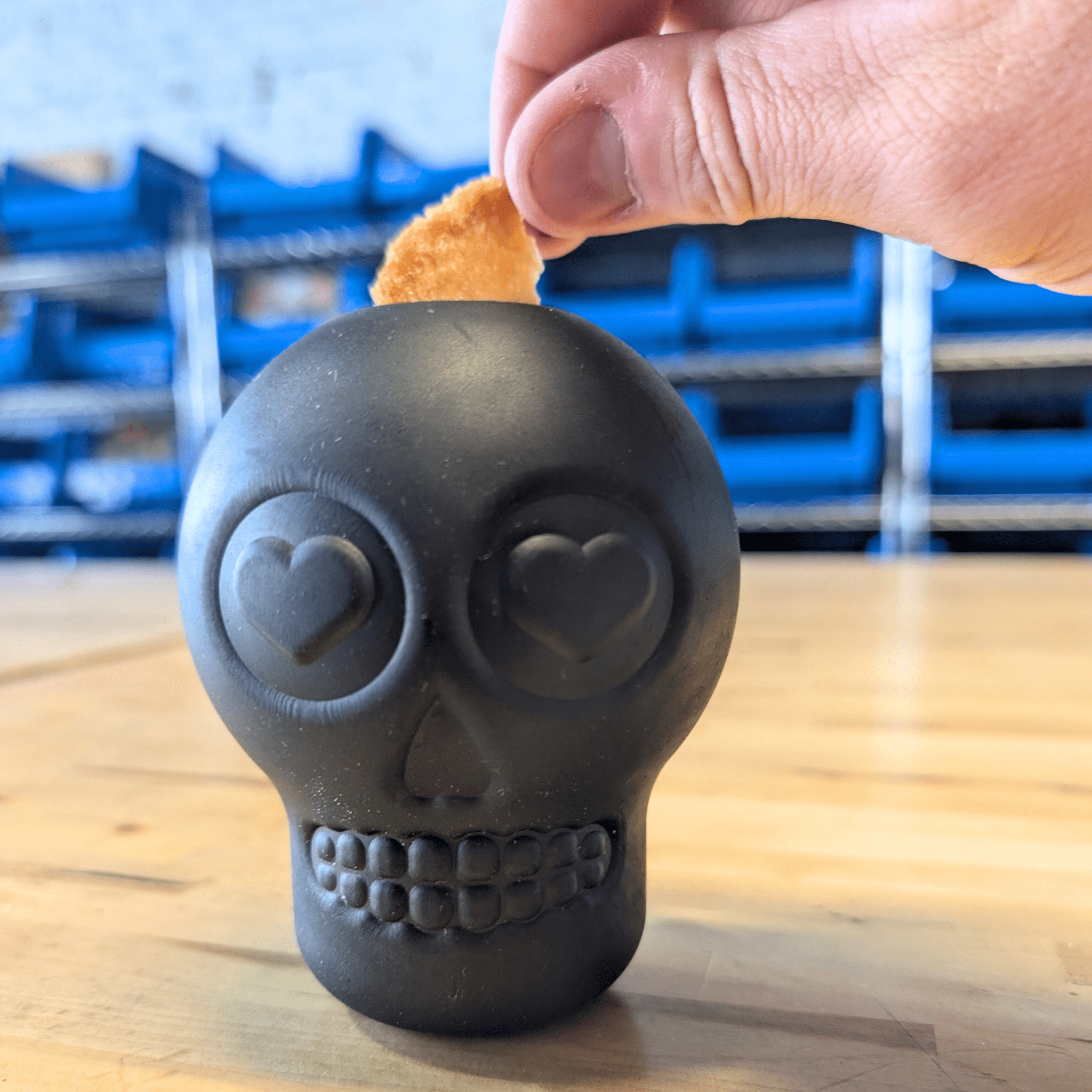 Farmto-pettreats Dog Treat Dispenser Black skull-shaped with heart eyes on a wooden surface a hand putting in a chip.