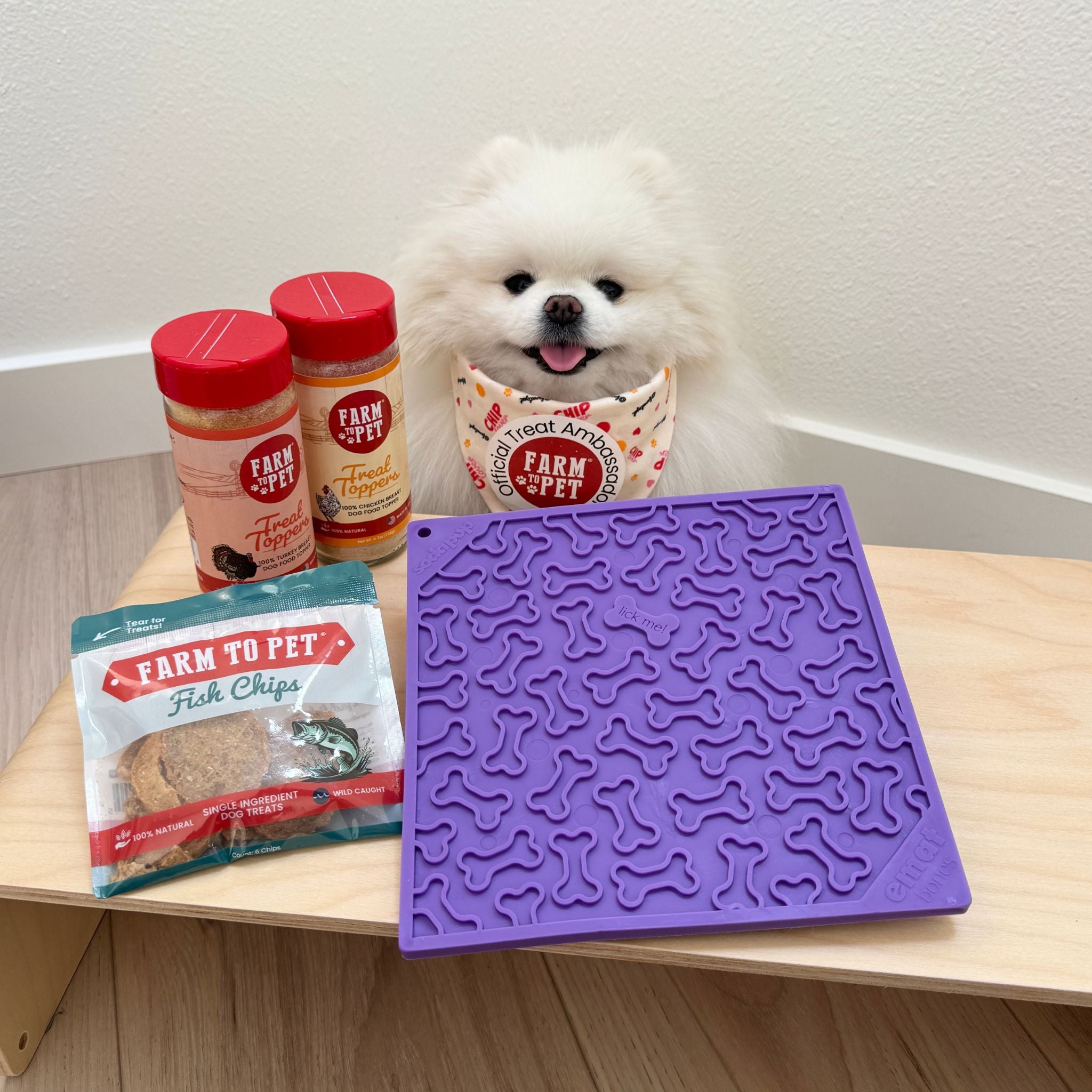 Farmto-pettreats purple dog bone lick mat benefits with a white dog wearing a bandana and Farmto-pettreats Fish Chips and Treat Toppers on a table.