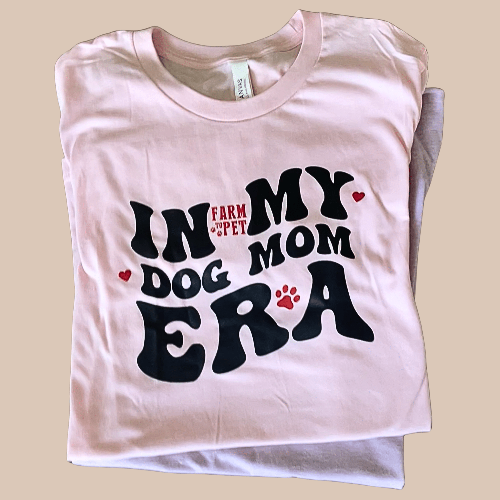 A folded Farmto-pettreats T-shirt with 'In My Dog Mom Era' text on a beige background.