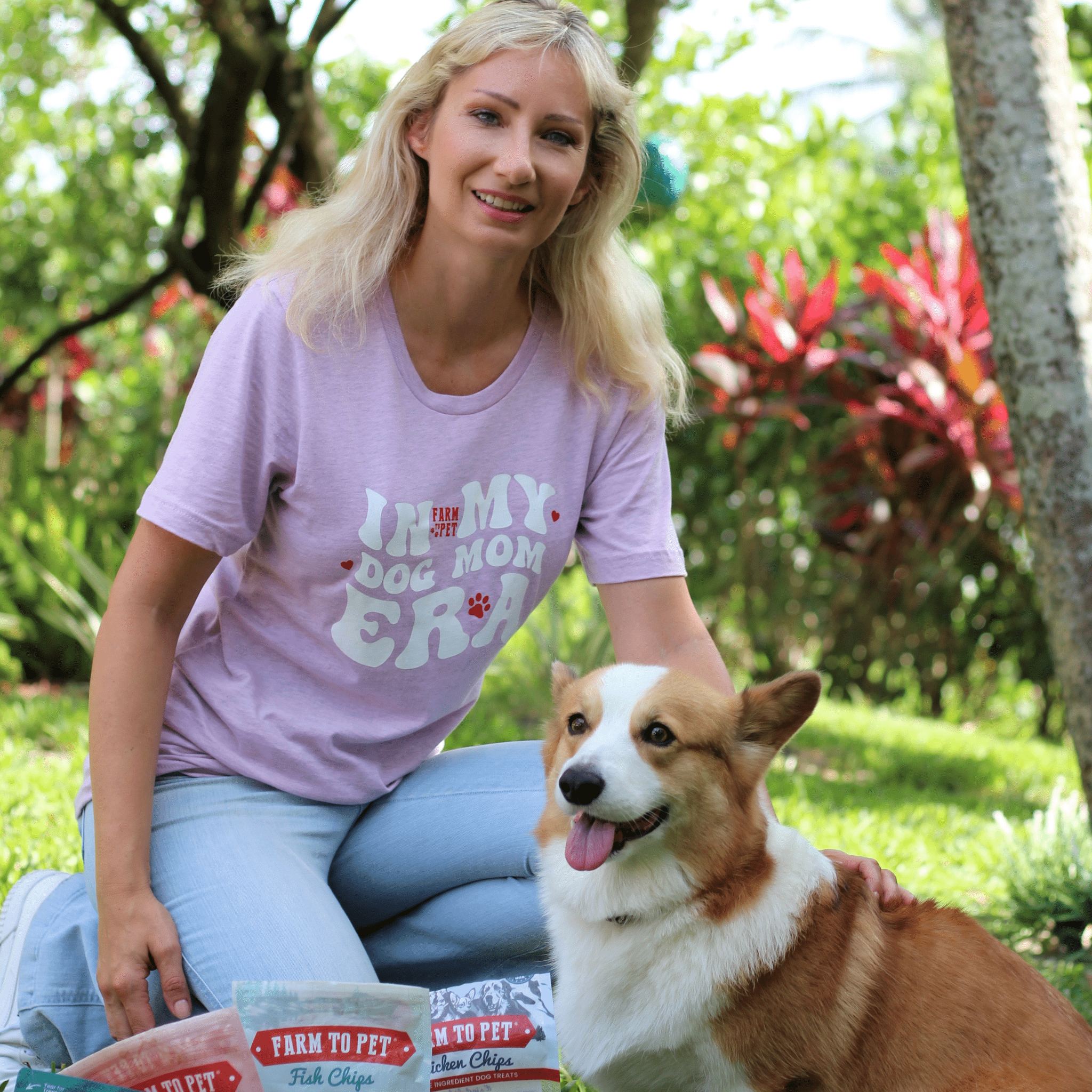 A woman wearing a purple Farmto-pettreats  t-shirt with a dog-themed design, sitting outdoors with a corgi.