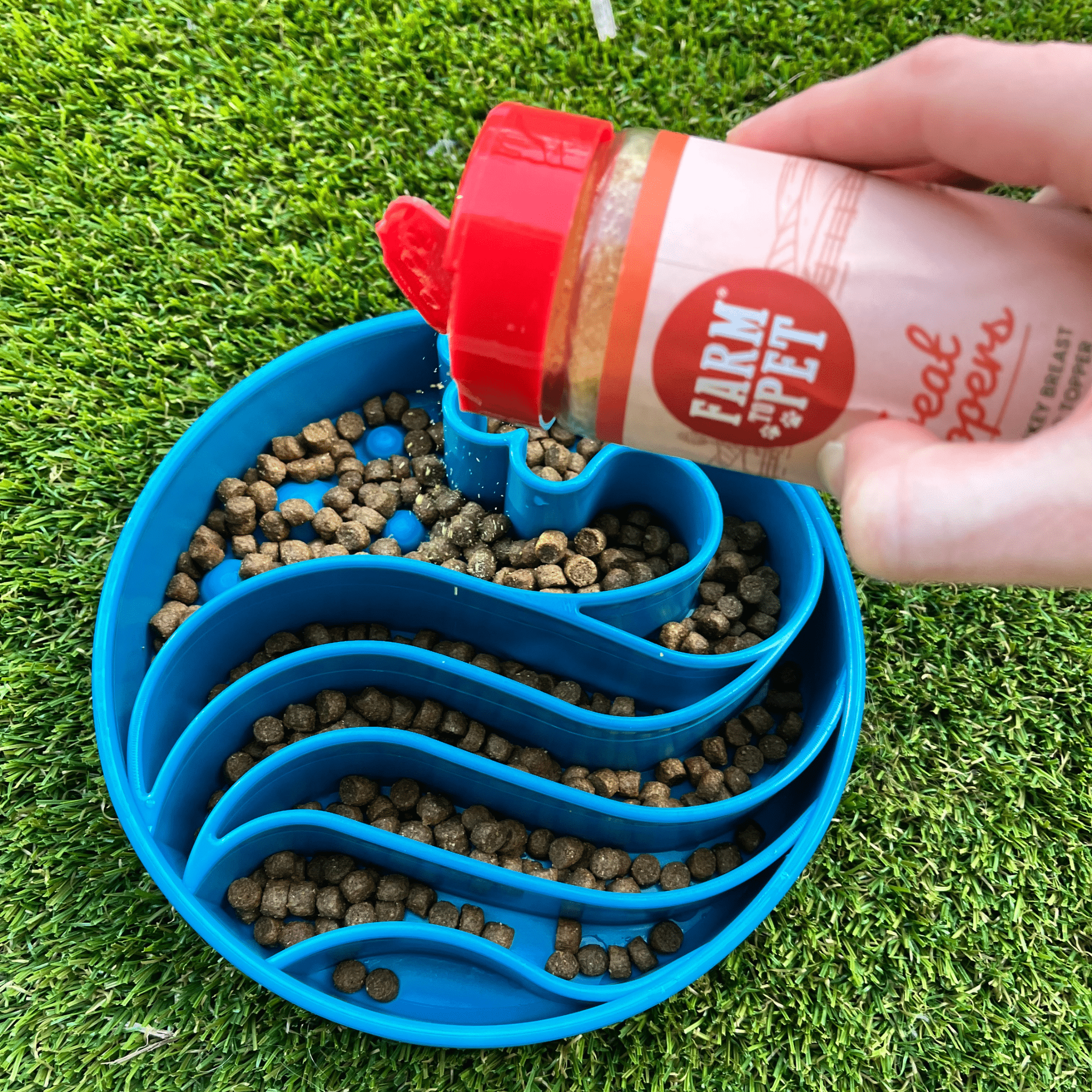 Hand pouring a product from Farmto-pettreats into a blue pet feeder filled with kibble on grass.